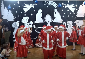 Na tle bożonarodzeniowej dekoracji dzieci w strojach Mikołajów wykonująMikołajowy Taniec. W środku tańczą chłopcy, po bokach tańczą dziewczynki.